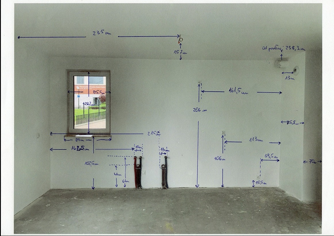Ściana z naniesionymi odręcznymi pomiarami i oznaczeniami instalacji elektrycznej, widoczne kable. Surowy stan pomieszczenia przed pracami wykończeniowymi.