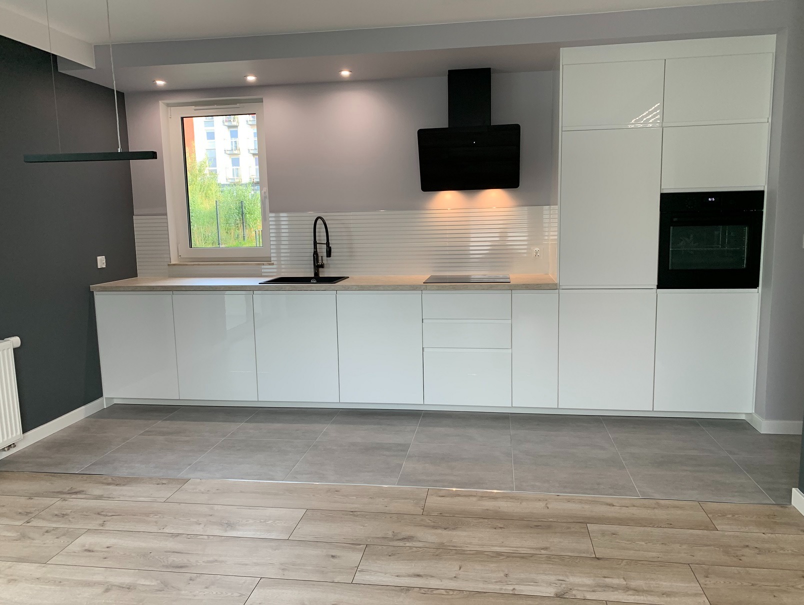 Nowoczesna, minimalistyczna kuchnia z białymi, błyszczącymi szafkami, czarną armaturą i okapem. Na podłodze szare płytki przechodzące w panele imitujące drewno.