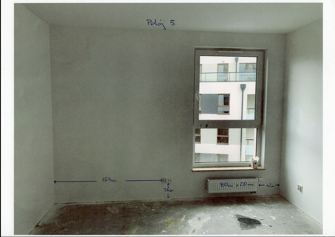 Pokój przed remontem: surowe ściany z odręcznymi notatkami wymiarów, okno z widokiem na budynek, widoczny grzejnik. Podłoga w stanie surowym, przygotowana do prac.