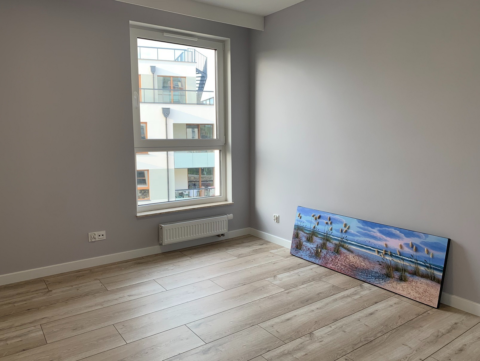 Jasne panele podłogowe w pokoju z szarą ścianą, oknem z widokiem na budynek i obrazem z motywem plaży opartym o ścianę. Widoczne gniazdka elektryczne i białe listwy przypodłogowe.