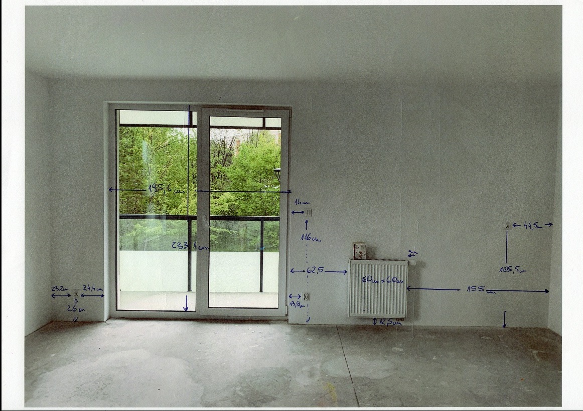 Pusty pokój z surowymi ścianami i betonową podłogą, na których naniesiono odręczne pomiary w centymetrach, okno balkonowe z widokiem na zieleń.