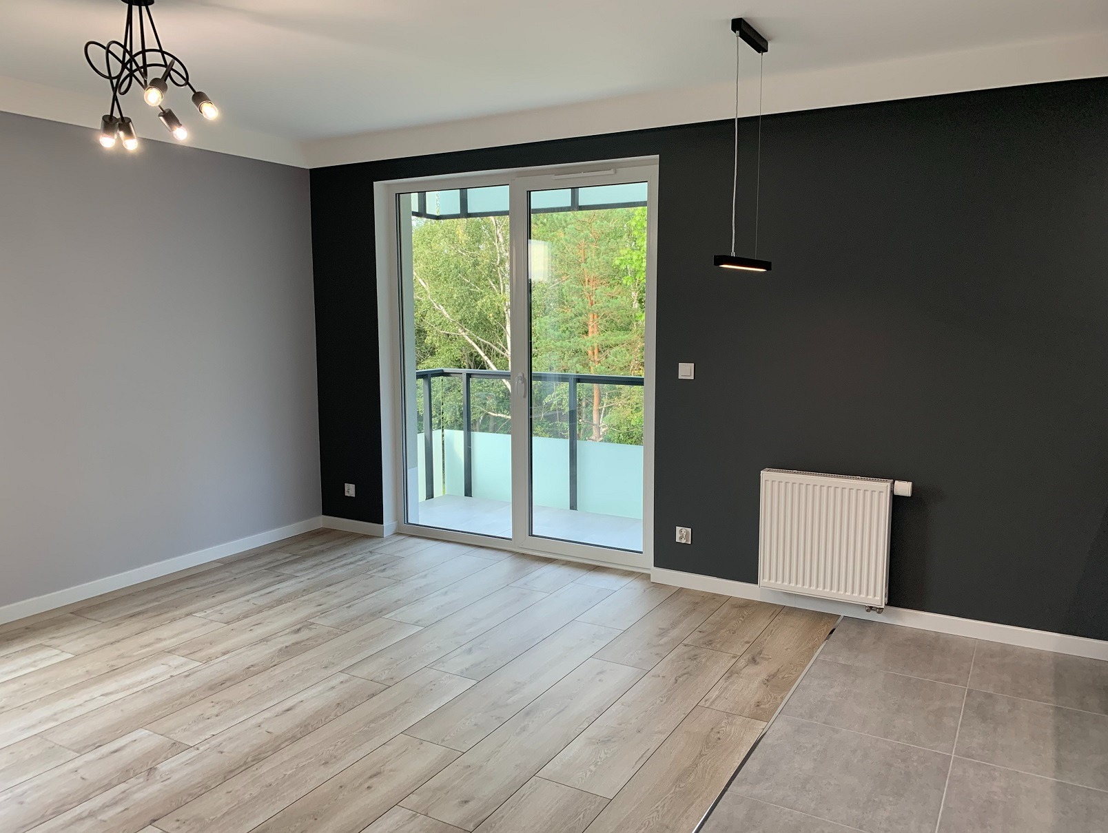 Jasne panele podłogowe w nowym pokoju z balkonem i grzejnikiem na ciemnej ścianie. Widok na zieleń za oknem. Minimalistyczna lampa sufitowa.