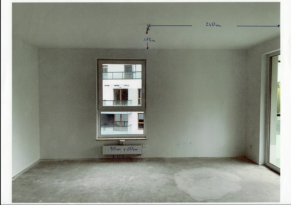 Surowe wnętrze z oknem i grzejnikiem, ściany przygotowane do malowania, widoczne odręczne notatki z wymiarami na ścianach i podłodze, stan deweloperski.