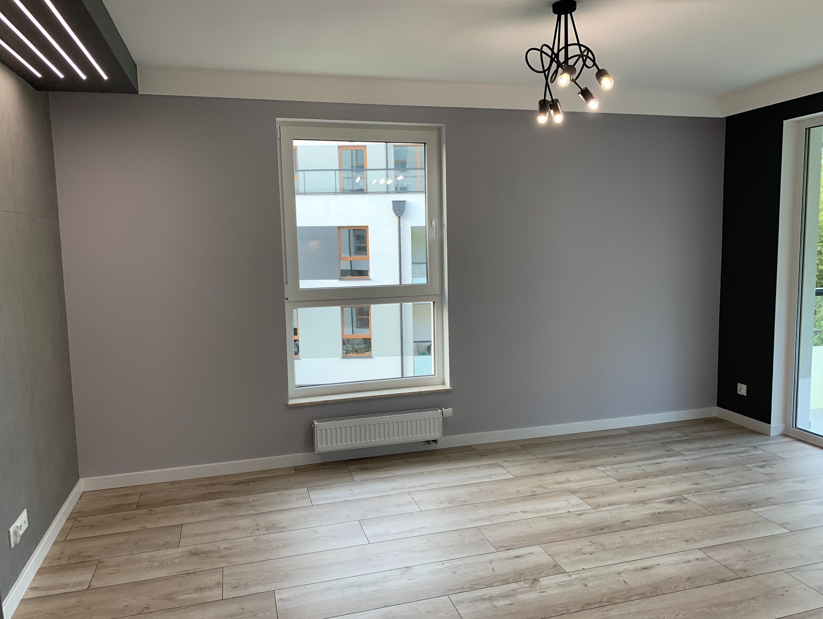 Jasne panele podłogowe w nowym mieszkaniu z szarą ścianą i oknem z widokiem na budynek. Nowoczesna lampa sufitowa. Minimalistyczne wykończenie wnętrza.