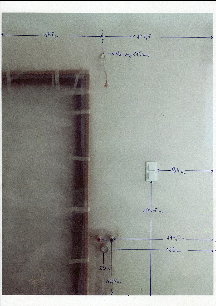 Ściana z naniesionymi odręcznie niebieskimi pomiarami i notatkami, widoczne gniazdko elektryczne, fragment ramy okiennej zabezpieczonej folią. Przygotowanie do prac wykończeniowych.
