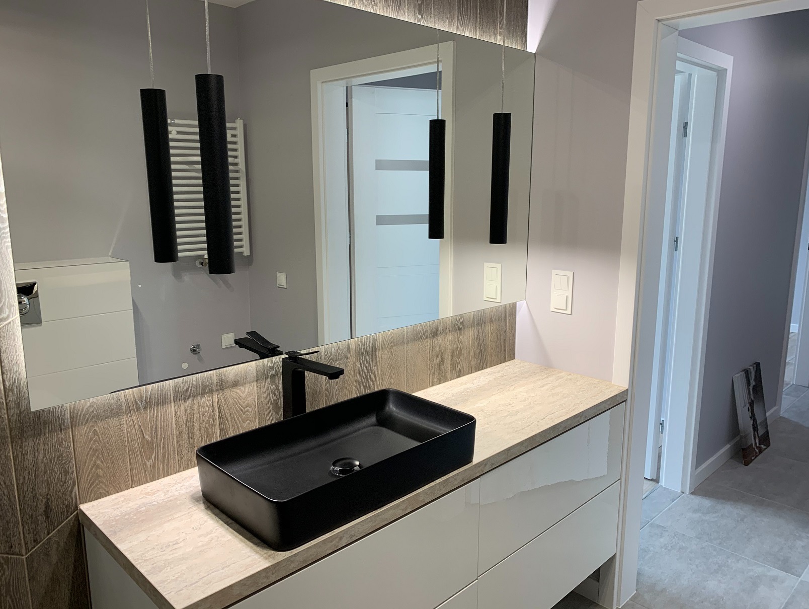 Nowoczesna łazienka z czarną, prostokątną umywalką nablatową, minimalistyczną armaturą i podświetlanym lustrem. Widoczne wiszące lampy i płytki imitujące drewno.
