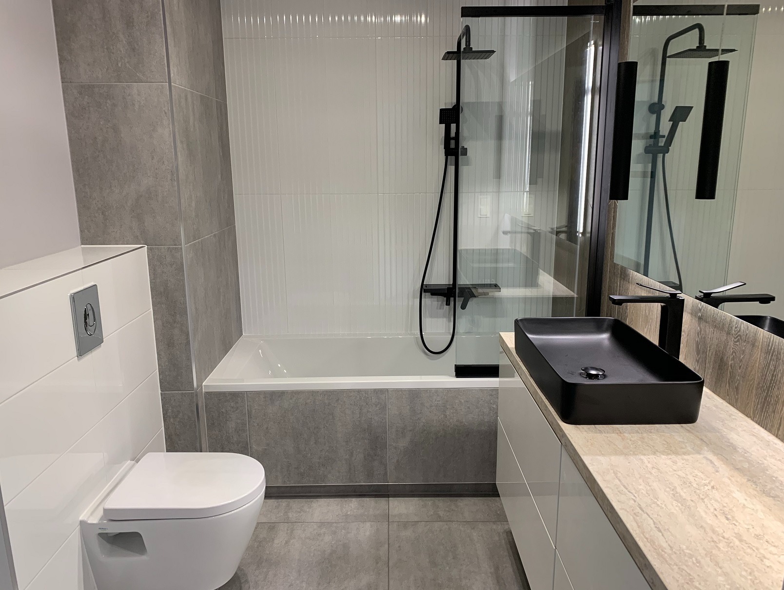 Nowoczesna łazienka z wanną obudowaną szarą płytką, białą armaturą i czarną, prostokątną umywalką nablatową. Minimalistyczny design, szaro-biała kolorystyka.