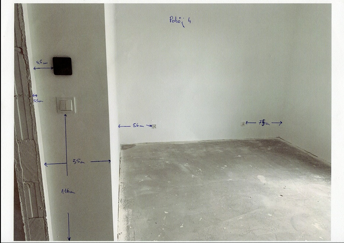 Surowy pokój z zaznaczonymi wymiarami na ścianach i podłodze, widoczne gniazdka elektryczne i włączniki. Ściany białe, podłoga betonowa. Widoczne ślady po remoncie.
