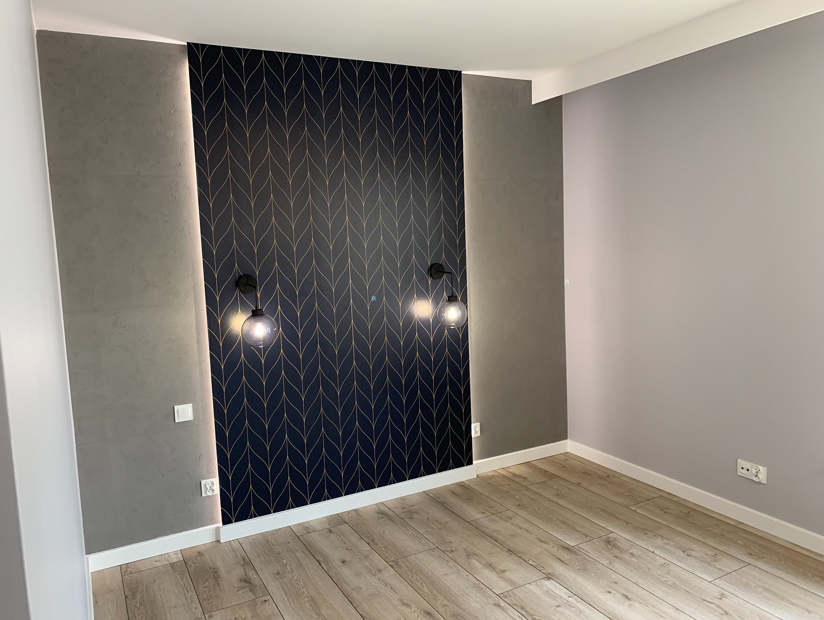 Pokój po metamorfozie: ściana z tapetą w geometryczny wzór, oświetlona dwoma lampami. Obok ściana w odcieniach szarości i podłoga z paneli w ciepłym odcieniu drewna.
