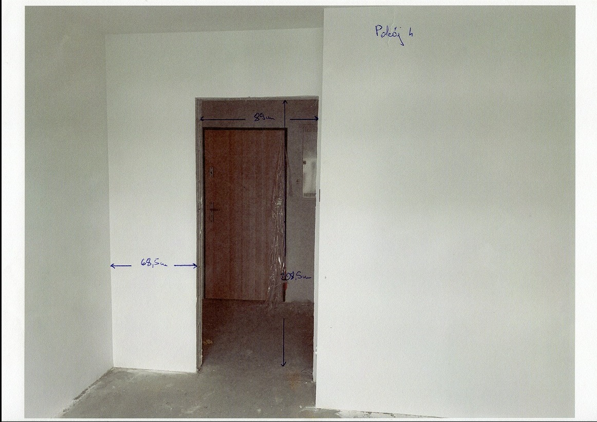 Surowy stan pokoju z zaznaczonymi wymiarami ścian i otworu drzwiowego, brązowe drzwi zabezpieczone folią. Notatki odręczne na ścianie.