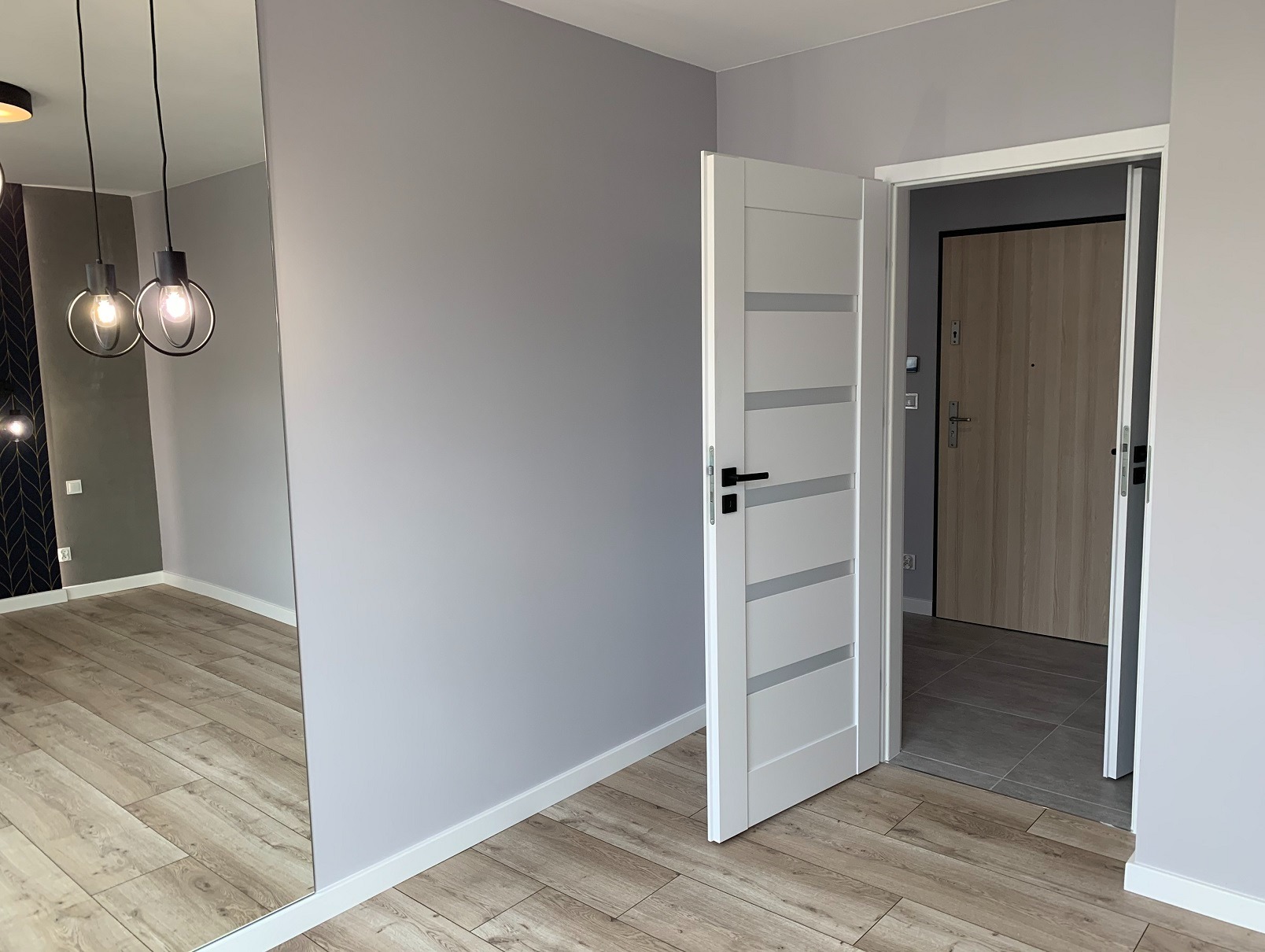 Szare ściany, jasne panele i białe drzwi z przeszkleniami w nowocześnie wykończonym wnętrzu. W lustrze odbija się designerska lampa i wzorzysta ściana.