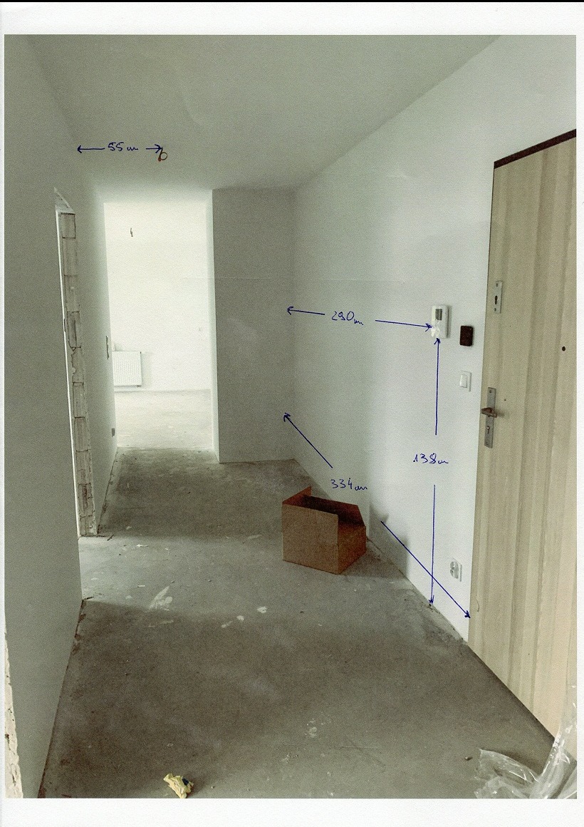 Surowy korytarz w trakcie prac: białe ściany z odręcznymi notatkami wymiarów, surowa podłoga, kartonowe pudło. Widok na dalsze pomieszczenia.