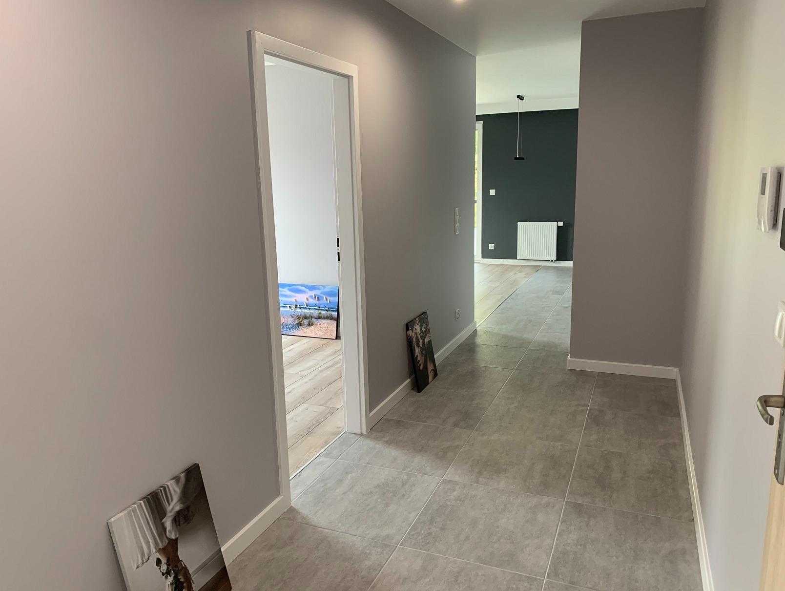 Pomalowane na szaro ściany korytarza z szarą podłogą z płytek. W tle widoczny pokój z ciemną ścianą i białym grzejnikiem. W przejściu obraz z motywem plaży.