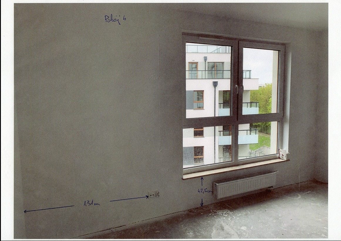 Surowy pokój z oknem i grzejnikiem, ściana z zaznaczonymi wymiarami i gniazdkiem. Widok na budynek z balkonami za oknem. Przygotowanie do prac wykończeniowych.