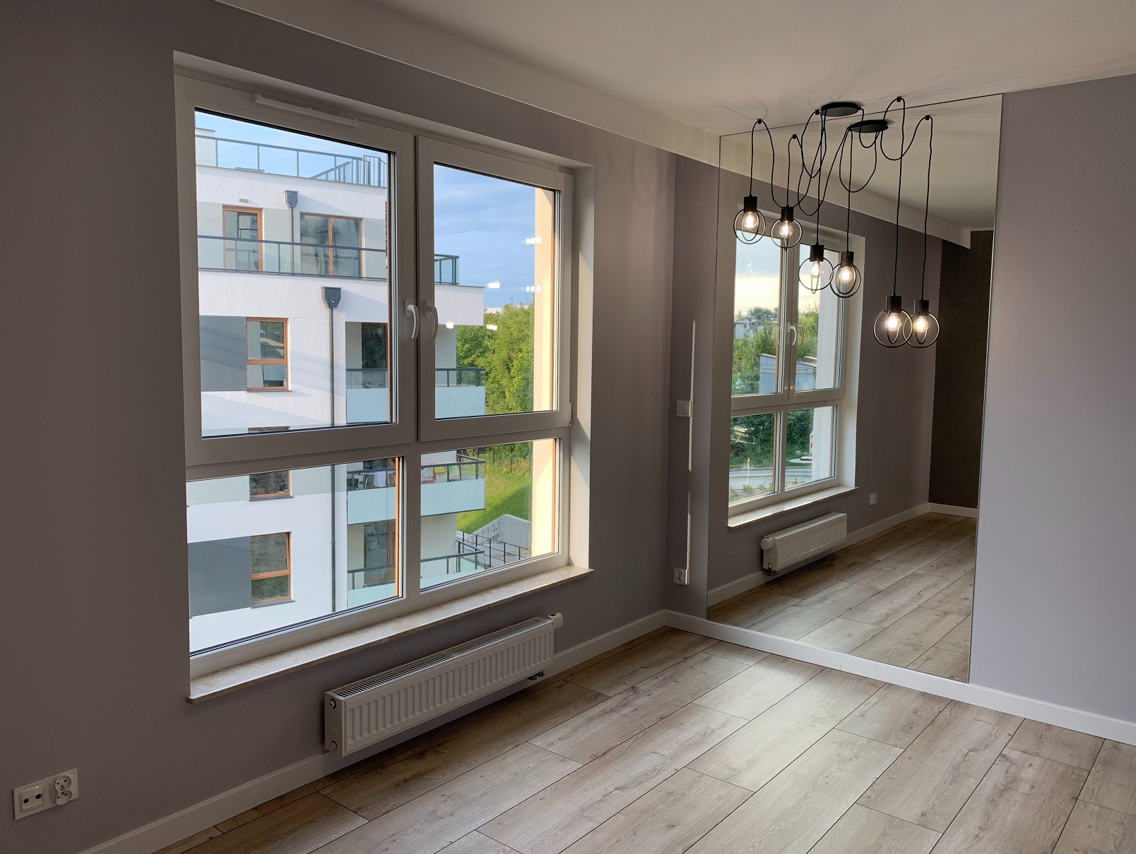 Jasne wnętrze po remoncie: parkiet, szare ściany, okna z widokiem na budynek, nowoczesna lampa wisząca i duże lustro na ścianie. Minimalistyczny design.