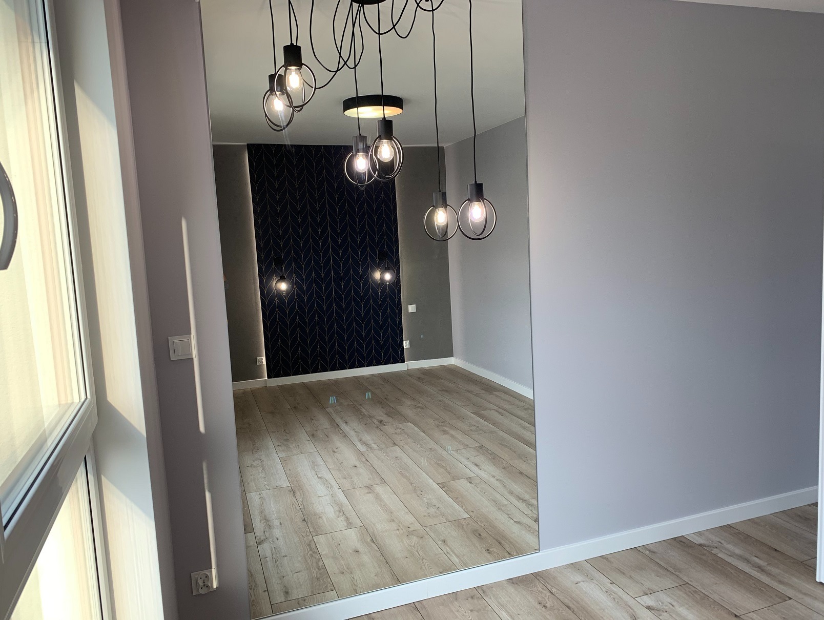 Nowoczesne wnętrze z lustrem: parkiet w jodełkę, designerska lampa wisząca i ściana akcentowa z geometrycznym wzorem. Widok z perspektywy okna.