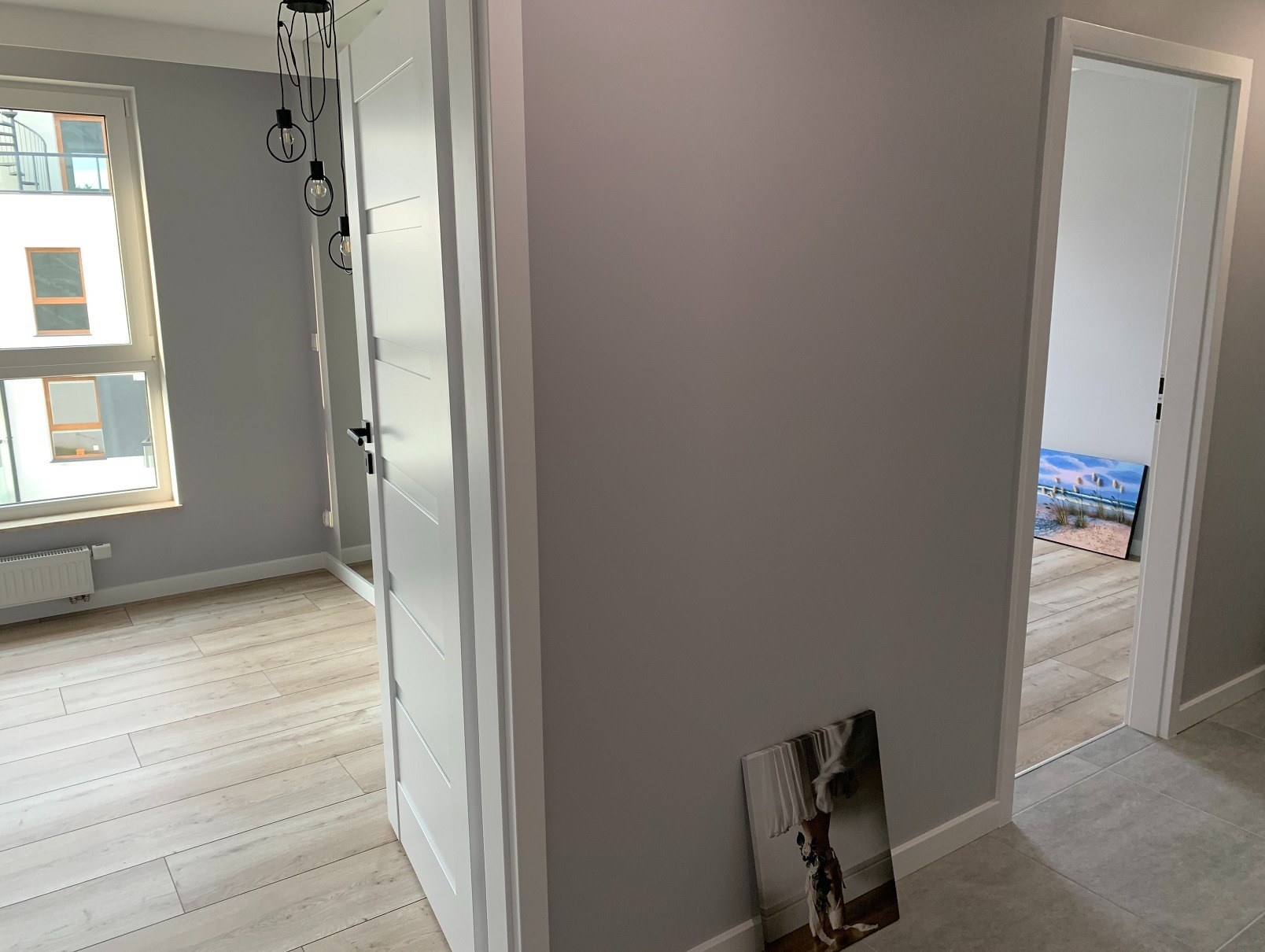 Jasne wnętrze mieszkania po remoncie: szare ściany, drewnopodobne panele, białe drzwi z czarną klamką, widok na okno i obraz na ścianie. Minimalistyczny design.