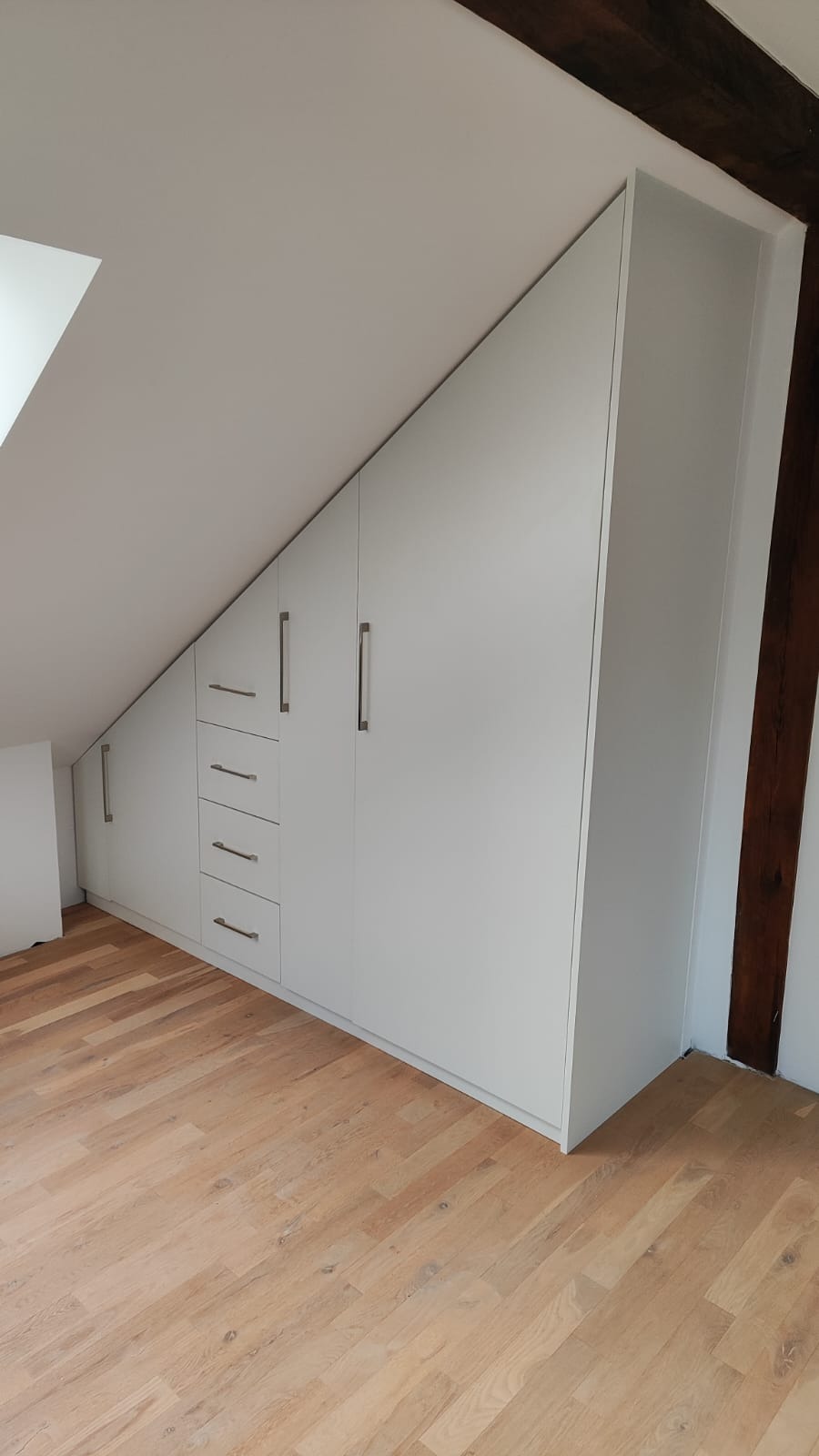 Biała szafa wnękowa na poddaszu ze skośnym sufitem, z drewnianą belką konstrukcyjną i czterema szufladami. Jasna podłoga z paneli.