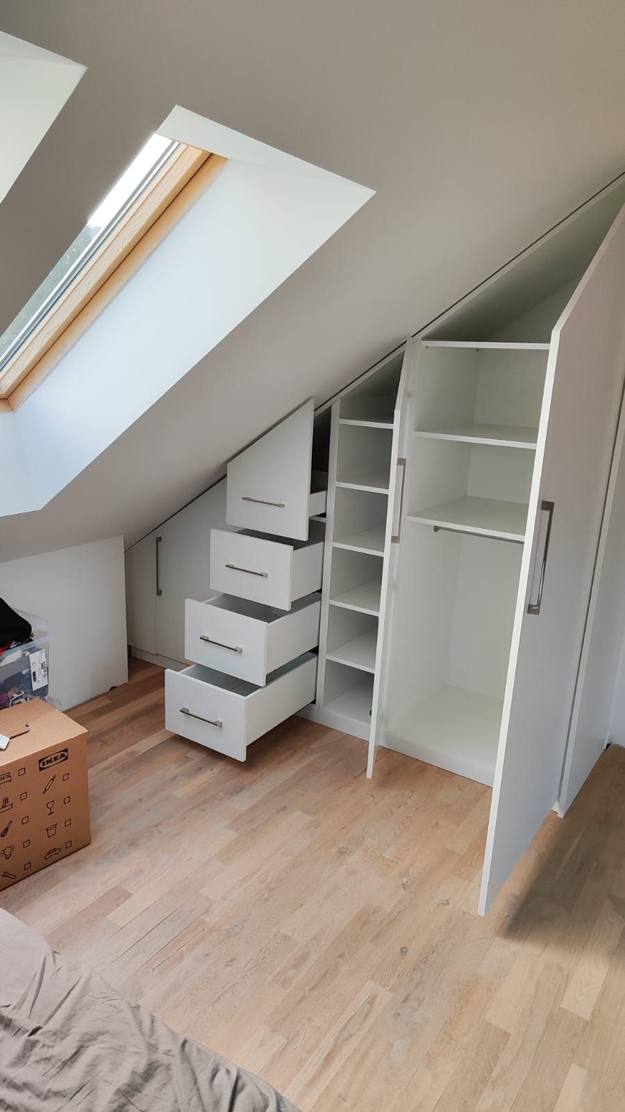 Biała szafa wnękowa ze schodkowymi szufladami, dopasowana do skosu poddasza, z otwartymi drzwiami ukazującymi półki i drążek na ubrania. Widok z dołu.