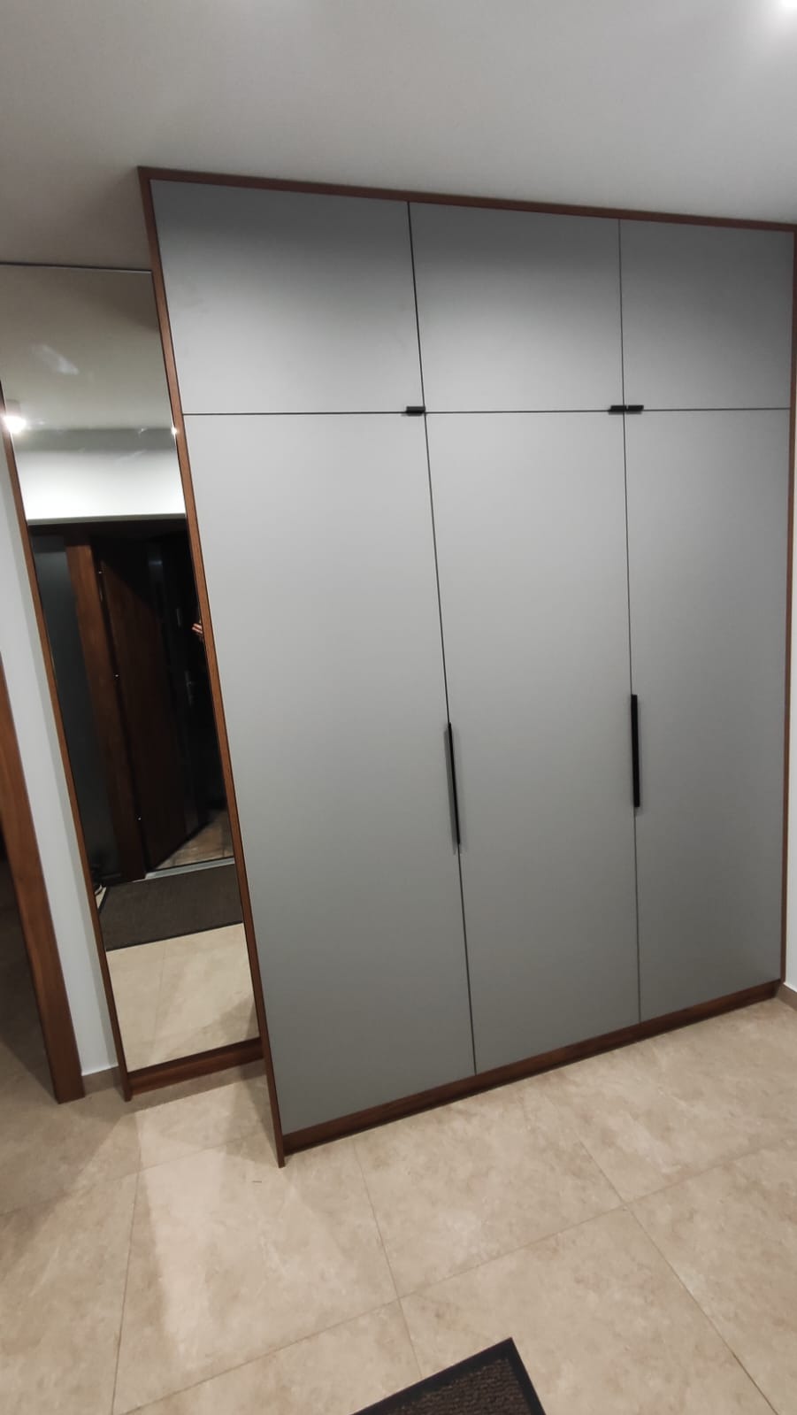 Szara szafa wnękowa z ciemnymi uchwytami, obok lustro w drewnianej ramie. Widoczna podłoga z płytek i fragment dywanika. Minimalistyczny design.