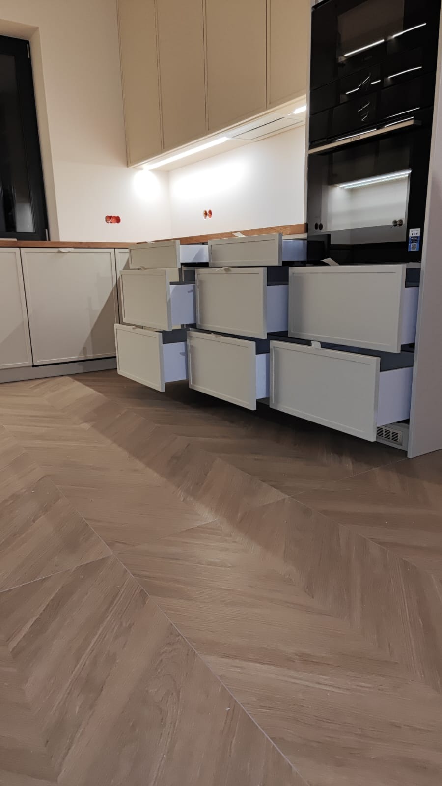 Jasna kuchnia na wymiar z otwartymi szufladami, ukazująca system organizacji przestrzeni. Parkiet ułożony w jodełkę. Widoczne oświetlenie podszafkowe i zabudowany piekarnik.