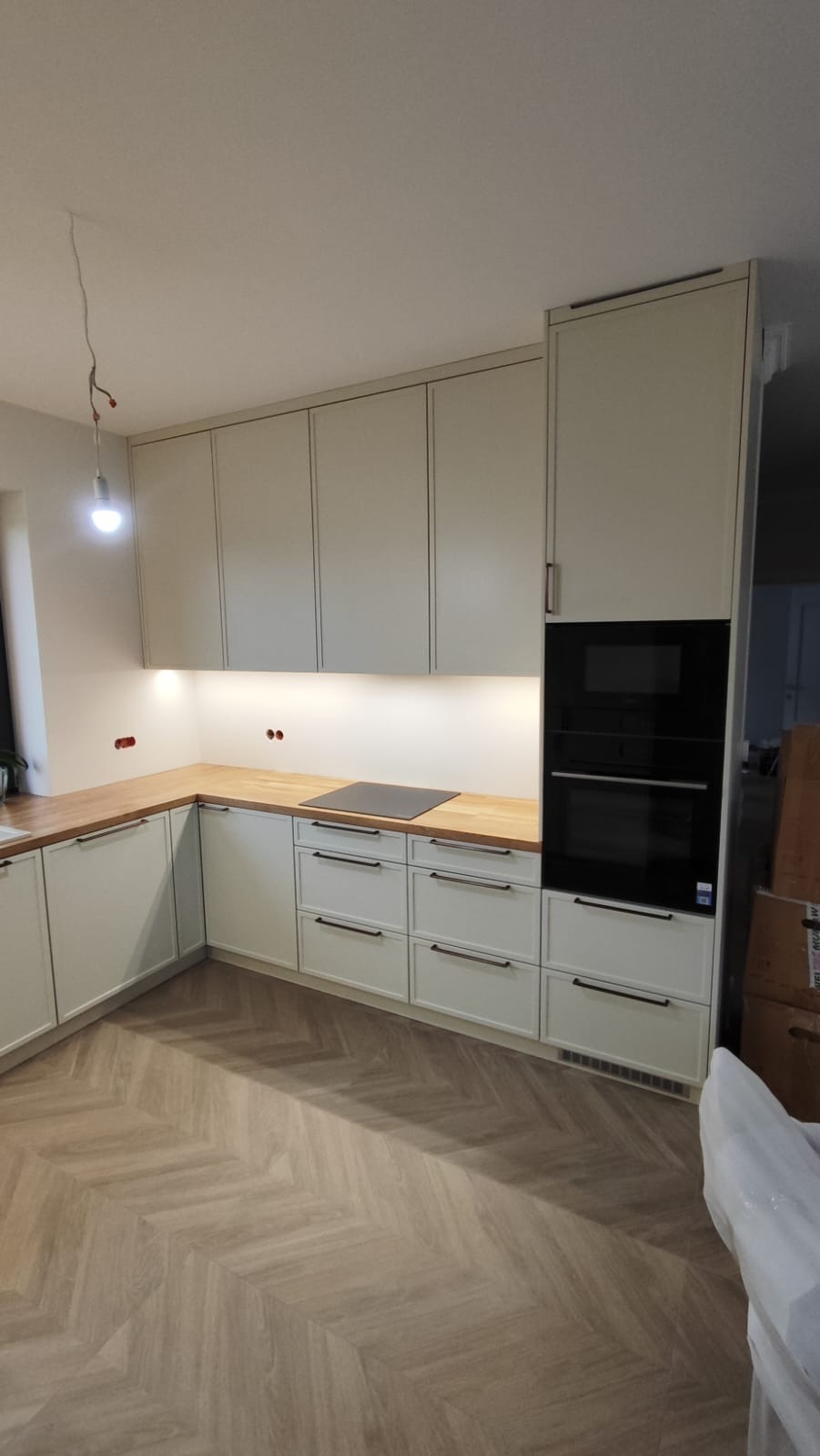 Jasna kuchnia z drewnianym blatem i czarnymi uchwytami, oświetlona listwą LED, z parkietem w jodełkę. Widoczny piekarnik w zabudowie i płyta indukcyjna.