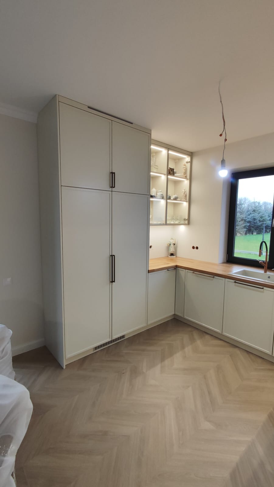 Jasna kuchnia z wysoką szafą i oświetloną witryną. Blat z drewna, podłoga w jodełkę. Nowoczesne, minimalistyczne wykończenie. Widok na zieleń za oknem.