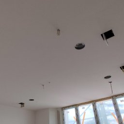 OKSANA KASHUBA OKSI-BUD - Biały sufit po szpachlowaniu z otworami na oświetlenie i wystającymi kablami, okno w tle. Prace wykończeniowe w nowym mieszkaniu.