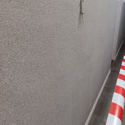 OKSANA KASHUBA OKSI-BUD - Ściana po szpachlowaniu i malowaniu z widoczną fakturą, zabezpieczona taśmą ostrzegawczą. Dwa wkręty w ścianie. Fragment korytarza w tle.