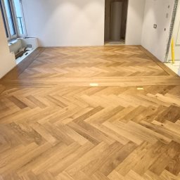 OKSANA KASHUBA OKSI-BUD - Ułożona parkietowa podłoga w jodełkę w jasnym, przestronnym wnętrzu. Widoczne detale wykończenia przy ścianach i oknie. W tle białe ściany i otwór drzwiowy.