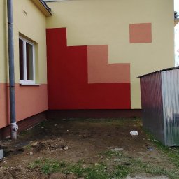 OKSANA KASHUBA OKSI-BUD - Elewacja budynku z geometrycznym wzorem w odcieniach czerwieni i beżu. Widoczne okno i fragment dachu. Teren wokół budynku częściowo zagospodarowany.