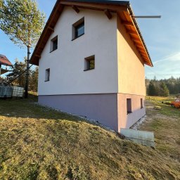 Otynkowany dom jednorodzinny z drewnianą konstrukcją dachu, widoczny z boku na tle zielonego wzgórza i błękitnego nieba w słoneczny dzień. Dolna część elewacji w innym kolorze.