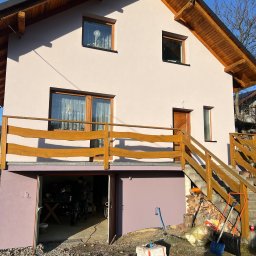 Aureo - Dwukondygnacyjny dom z drewnianą konstrukcją dachu i balustradą, świeżo otynkowany na jasny kolor, z otwartym garażem i narzędziami malarskimi na zewnątrz.