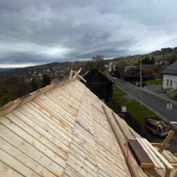 Konstrukcja dachu z desek w trakcie budowy, widok z góry na tle zabudowań i pochmurnego nieba. Widoczne elementy mocujące i narzędzia.