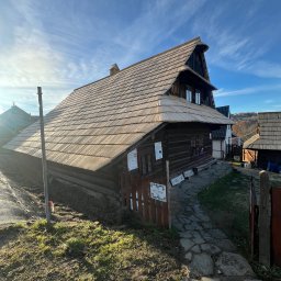 Drewniany dom w stylu góralskim z gontowym dachem, oświetlony słońcem na tle błękitnego nieba. Widoczna kamienna ścieżka i elementy krajobrazu.