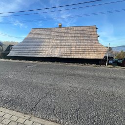 Drewniany dom z gontowym dachem w stylu regionalnym, widok z poziomu ulicy. Charakterystyczna architektura, błękitne niebo w tle.