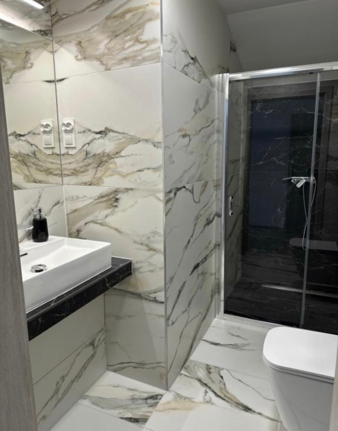 Elegancka łazienka z marmurowymi płytkami, kabiną prysznicową i umywalką nablatową. Nowoczesny design z akcentami czerni i bieli. Widoczne detale wykończenia.