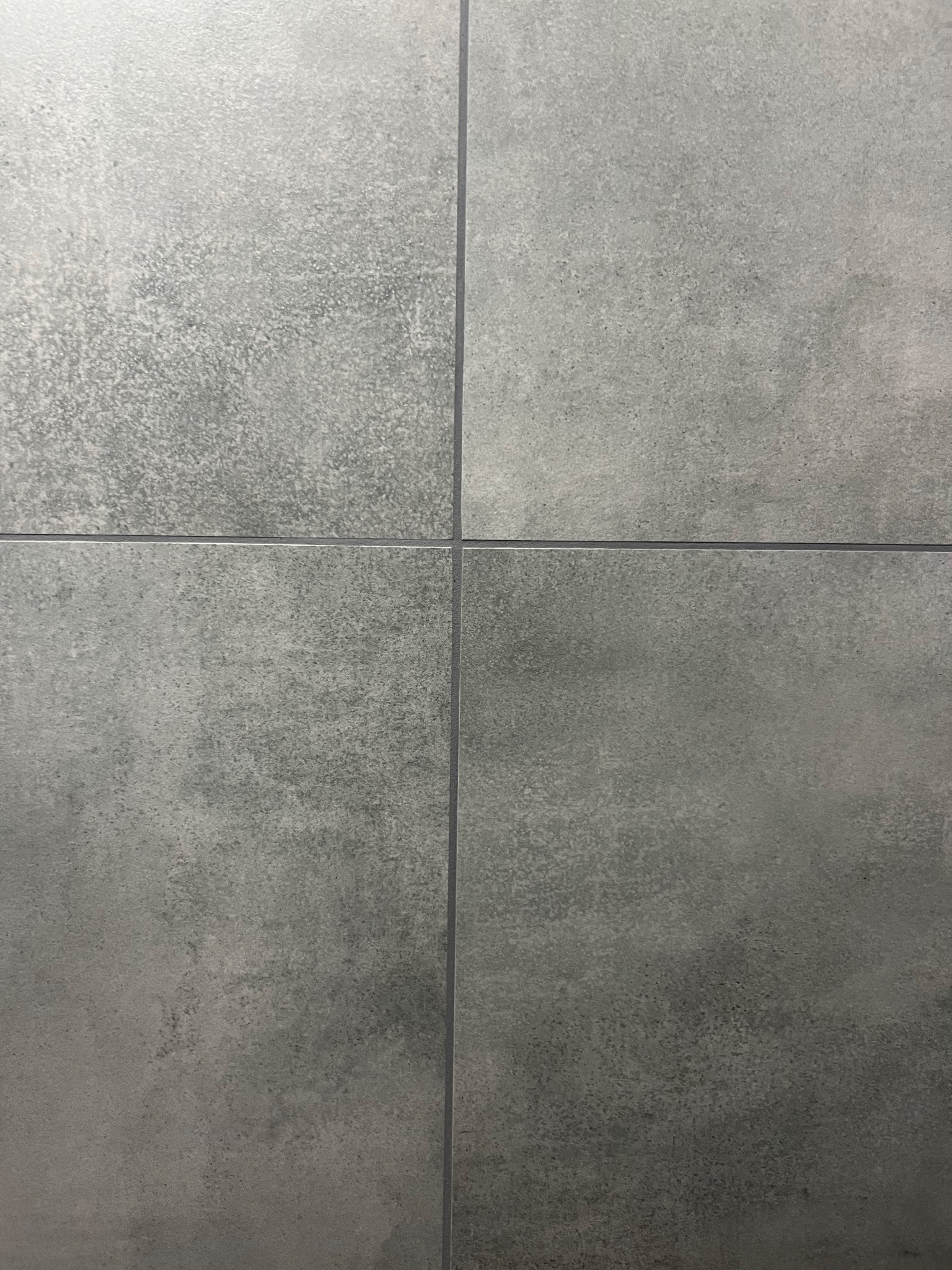 Szare, matowe płytki gresowe ułożone w kwadrat, z widocznymi fugami. Minimalistyczny wzór, imitujący beton, z delikatną teksturą i subtelnymi różnicami w odcieniach szarości.