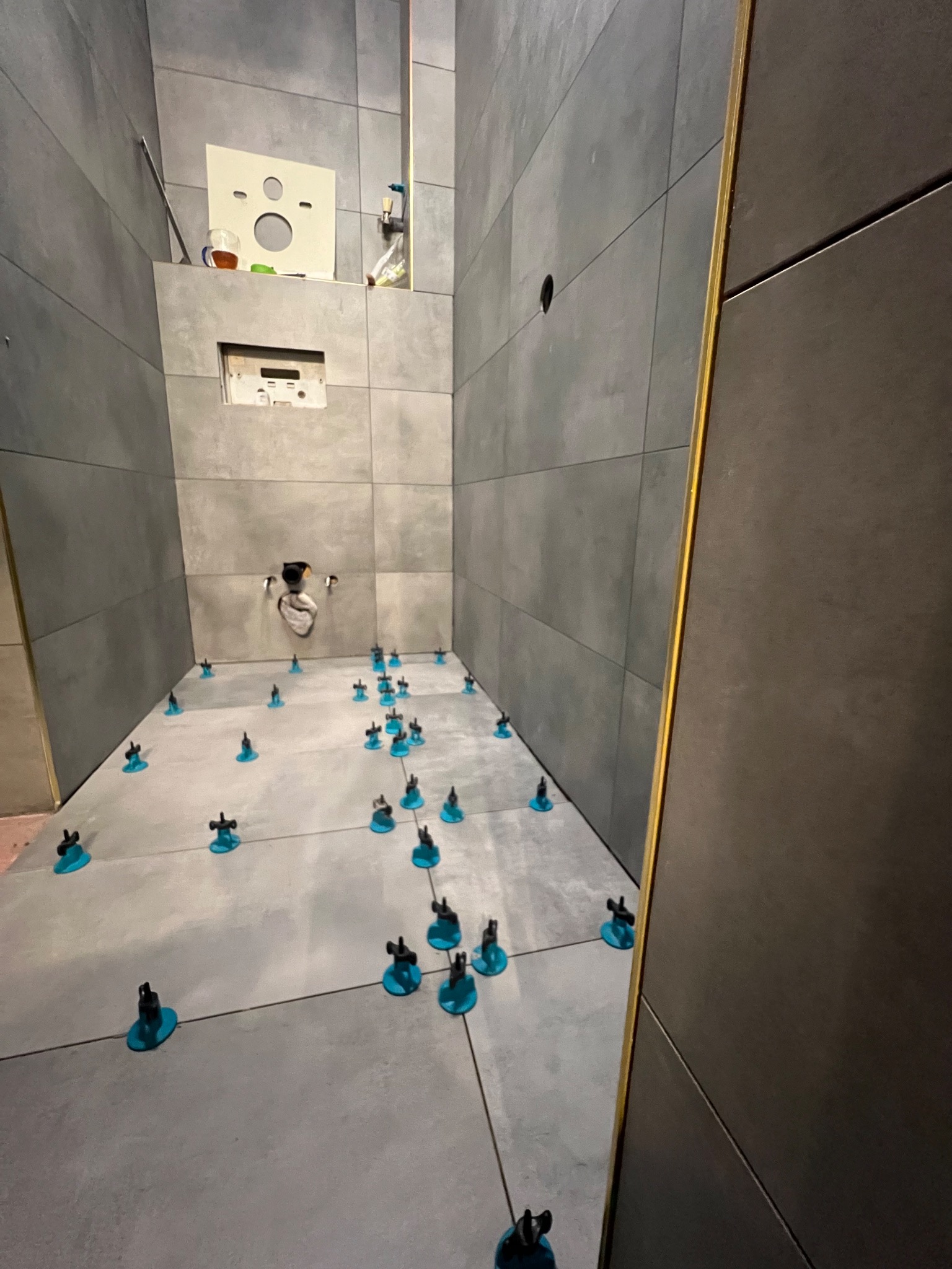 Szare płytki w łazience, wyrównane niebieskimi klipsami poziomującymi. Widoczne otwory na instalacje i fragment ściany z żółtą listwą wykończeniową.