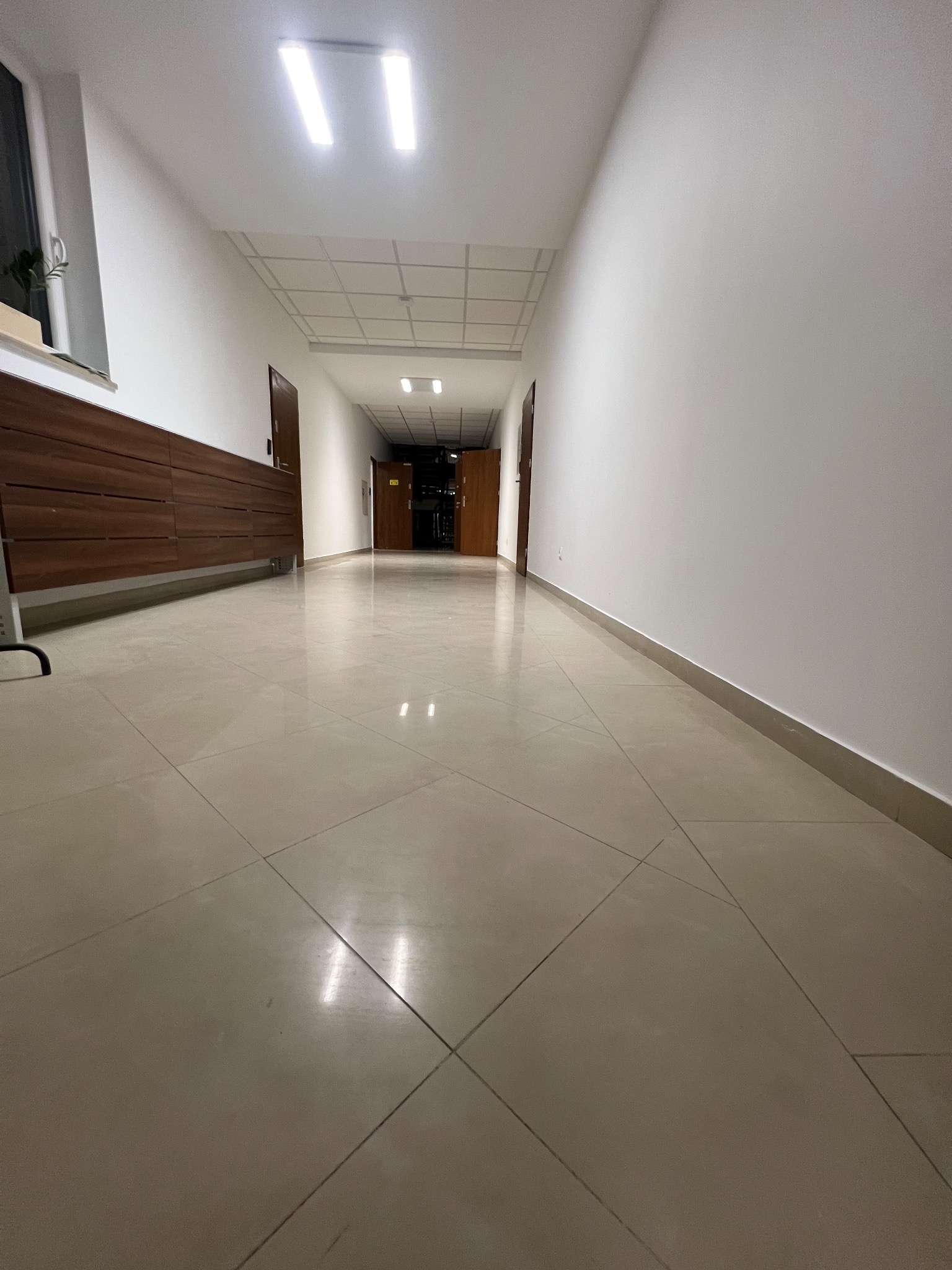 Długi, pusty korytarz biurowy z beżową podłogą z płytek i białymi ścianami. Widoczne drzwi po bokach i oświetlenie sufitowe. Perspektywa z dołu.