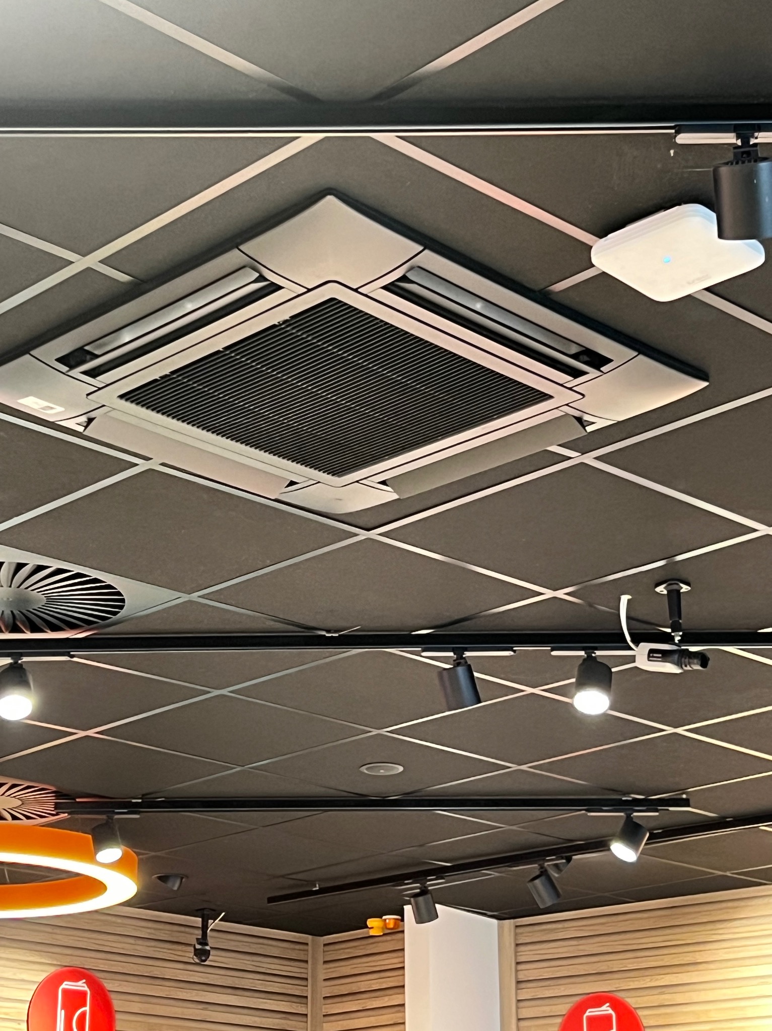 Kasetonowy sufit z wbudowaną klimatyzacją i oświetleniem punktowym. Widoczne kratki wentylacyjne i elementy instalacji. Ciemna kolorystyka sufitu z jasnymi liniami podziału.