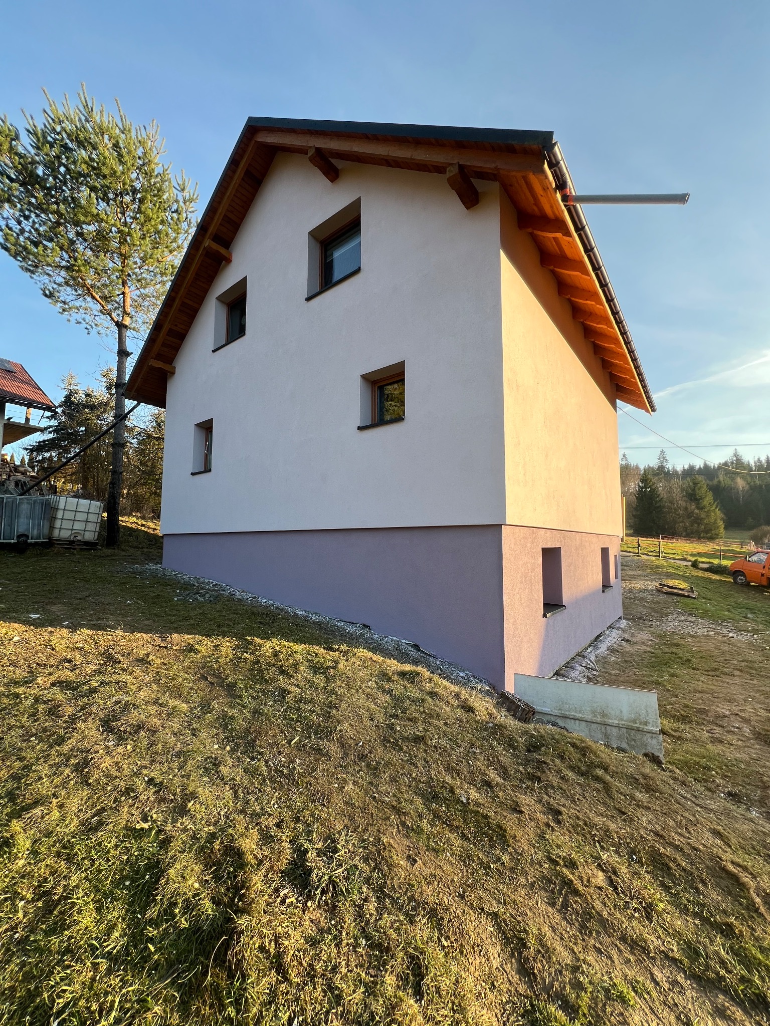 Otynkowany dom jednorodzinny z drewnianą konstrukcją dachu, widoczny z boku na tle zielonego wzgórza i błękitnego nieba w słoneczny dzień. Dolna część elewacji w innym kolorze.