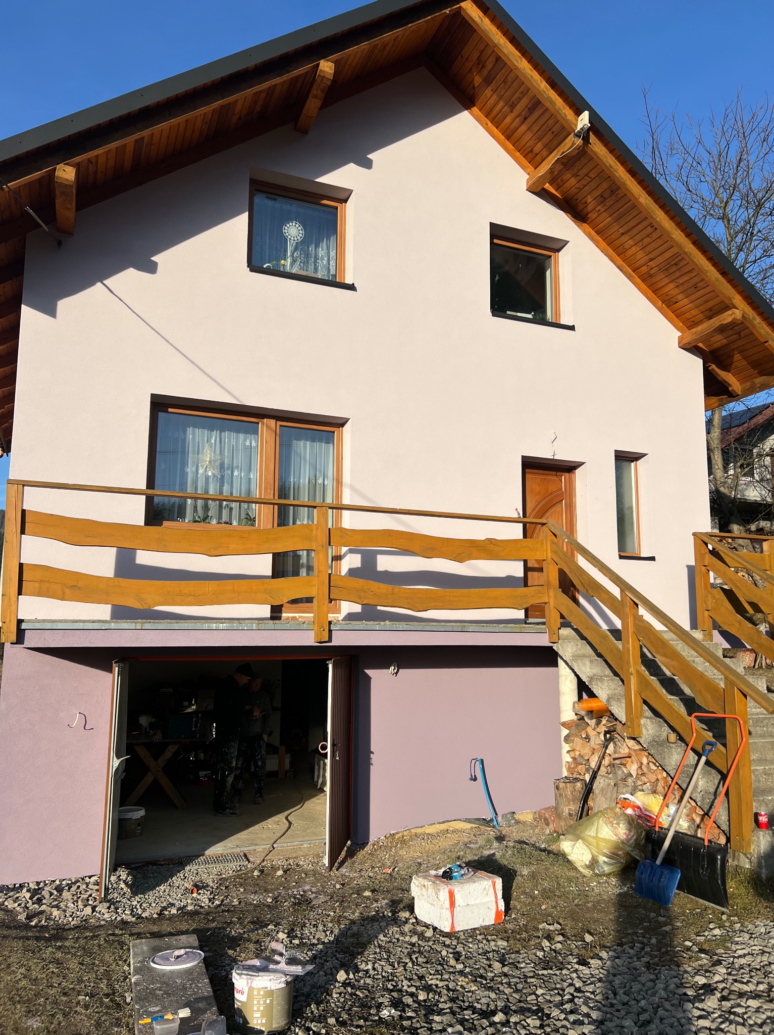 Dwukondygnacyjny dom z drewnianą konstrukcją dachu i balustradą, świeżo otynkowany na jasny kolor, z otwartym garażem i narzędziami malarskimi na zewnątrz.