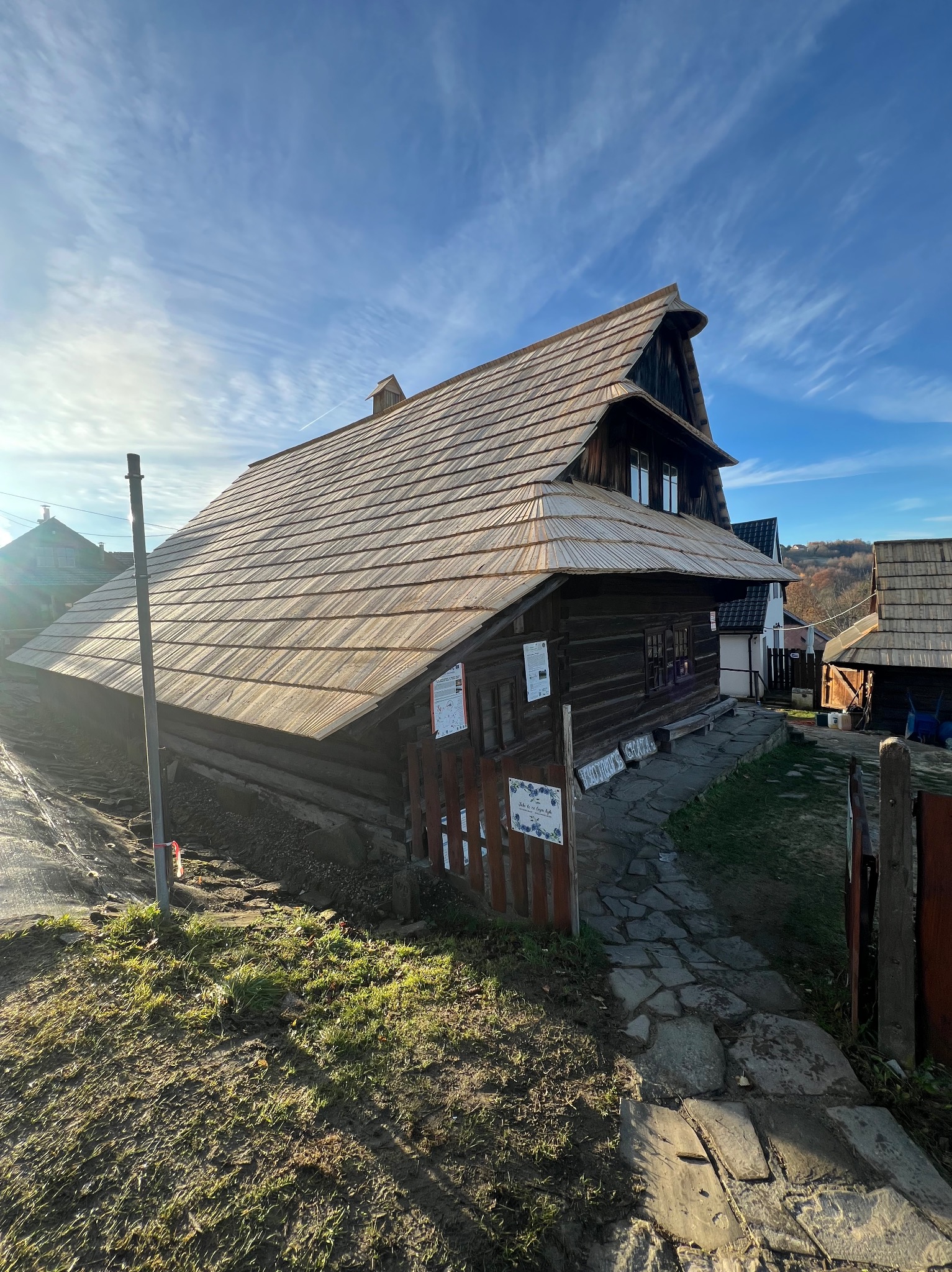 Drewniany dom w stylu góralskim z gontowym dachem, oświetlony słońcem na tle błękitnego nieba. Widoczna kamienna ścieżka i elementy krajobrazu.