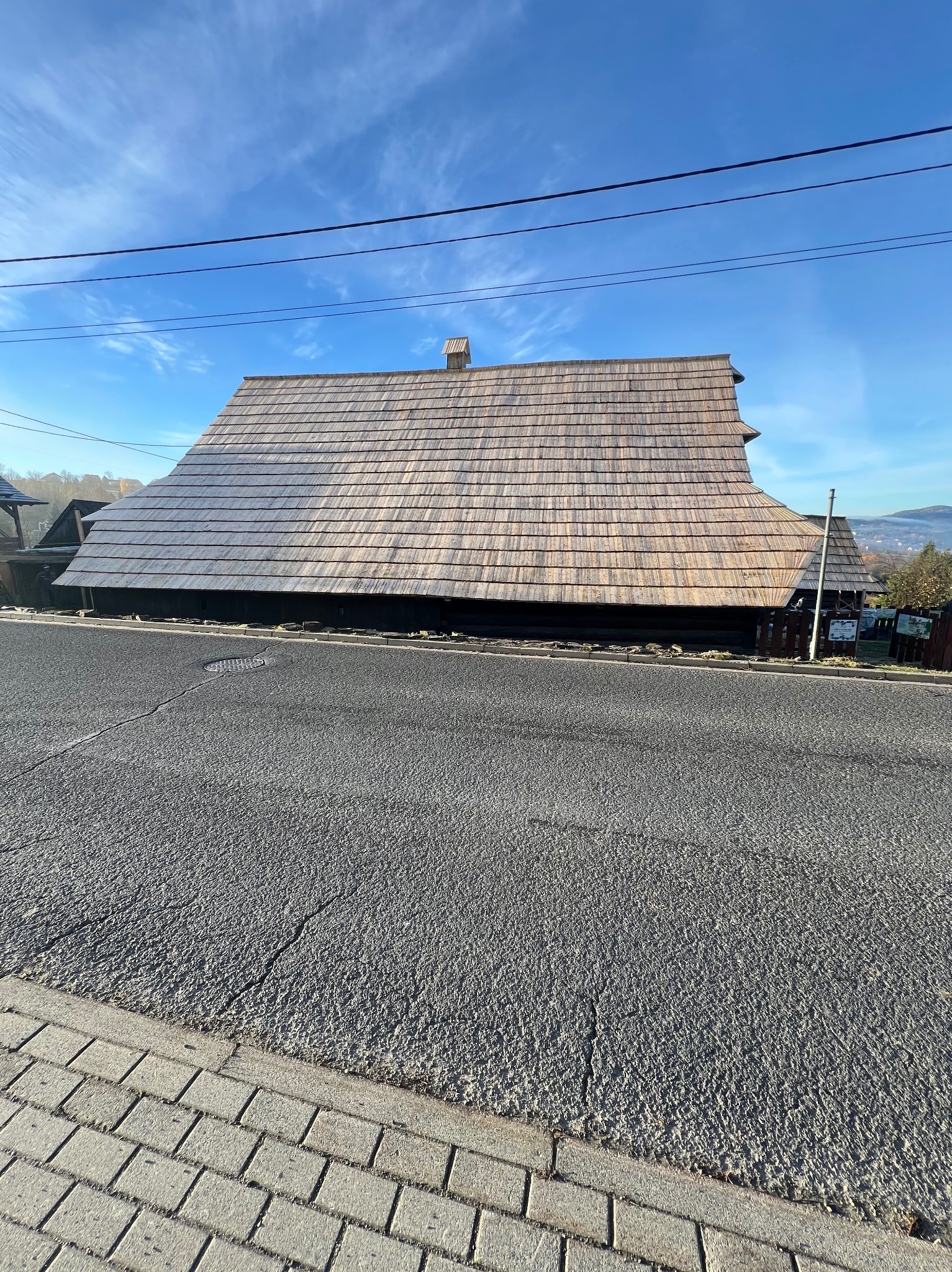 Drewniany dom z gontowym dachem w stylu regionalnym, widok z poziomu ulicy. Charakterystyczna architektura, błękitne niebo w tle.