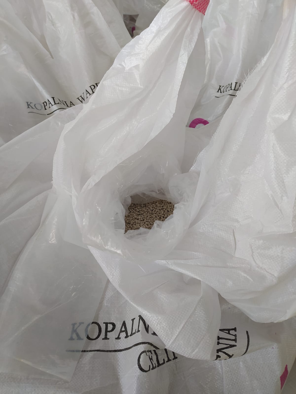 Otwarty worek nawozu granulowanego firmy Kopalnia Wapienia Cielętniki, widoczne szare granulki nawozu wewnątrz worka. W tle inne worki.