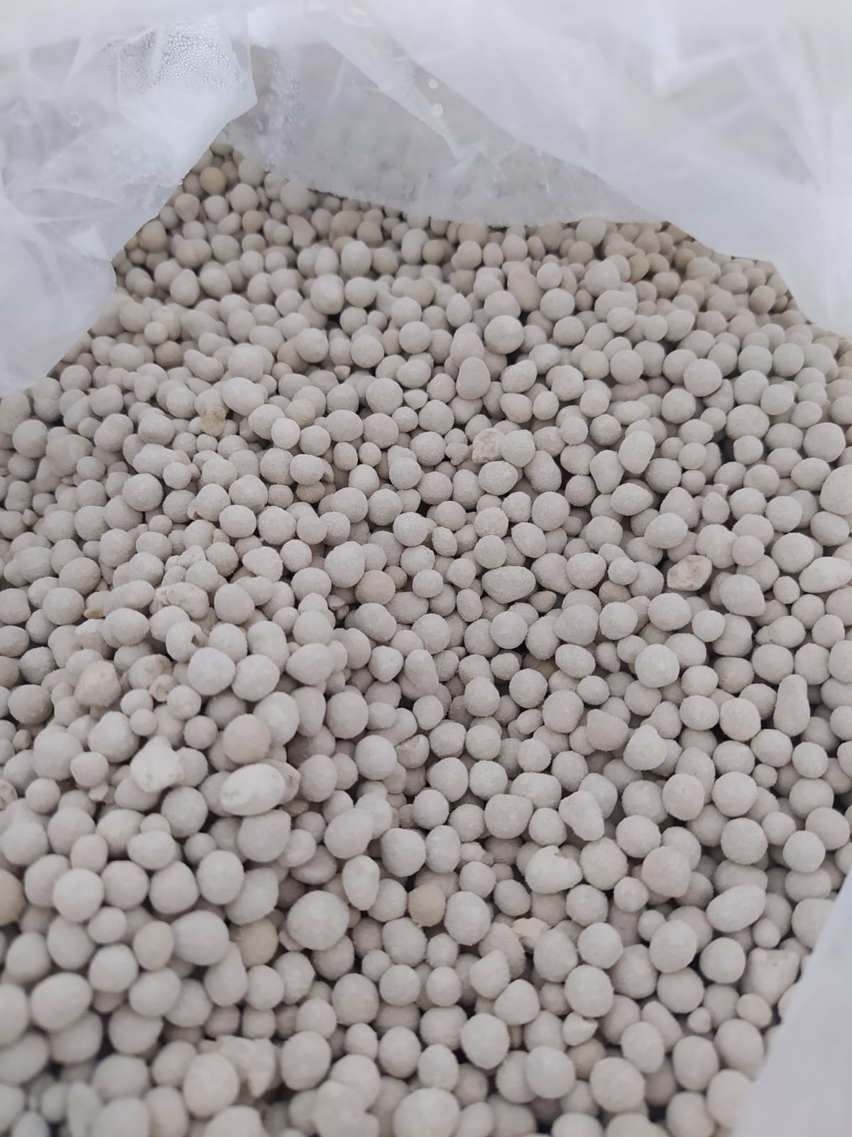 Jasnoszare granulki w przezroczystym worku, prawdopodobnie nawóz mineralny. Widoczne nierówności i zróżnicowanie wielkości granulek.