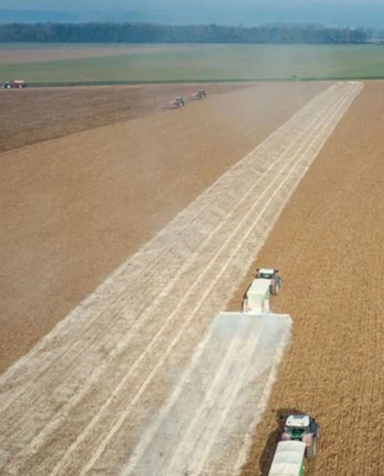 Rozległe pole z ciągnikami rozsiewającymi substancje, widok z góry. Ślady na polu wskazują na postęp prac. Horyzont zalesiony w tle.