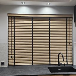 Świat Rolet i Żaluzji - Jasne, poziome przesłony okienne z czarnymi taśmami w nowoczesnej kuchni z drewnianymi szafkami i czarnym zlewem. Oświetlenie sufitowe.