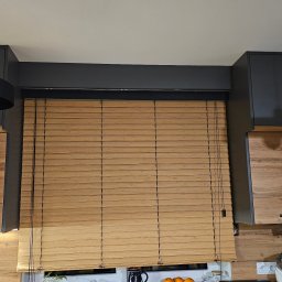 Świat Rolet i Żaluzji - Jasne, poziome lamele w oknie kuchennym, drewniane szafki z widocznym usłojeniem, czarne lampy wiszące i ciemnoszare szafki górne tworzą spójną całość.